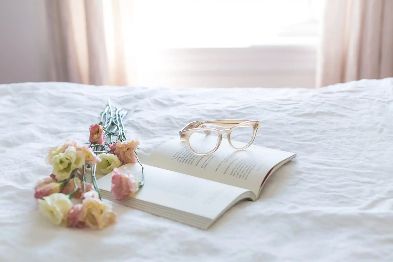 Open book with reading glasses and delicate flowers resting on white bedding.